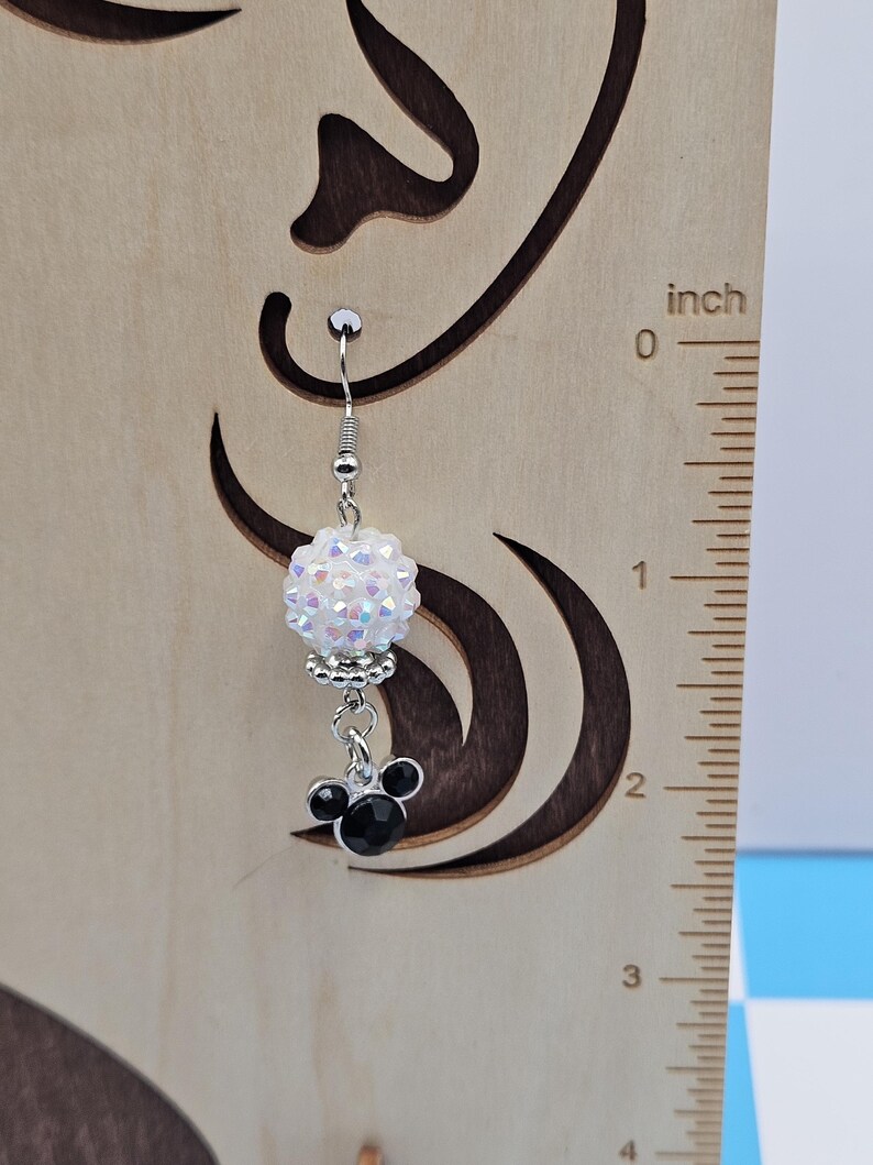 May include: A single silver earring with a white, iridescent, beaded ball and a black Mickey Mouse charm. The earring is hanging in front of a wooden background with a ruler showing inches.