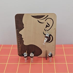 May include: A wooden display stand with a profile silhouette and a ruler in inches. A pair of ice cream sundae earrings are displayed on the stand, along with three miniature ice cream sundae figurines. The background is a pink grid pattern.