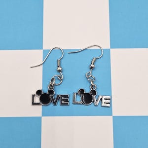 May include: Silver-toned dangle earrings. Each earring features the word "LOVE" with a black silhouette of a mouse head replacing the "O". The earrings hang from silver-colored fish hook ear wires.