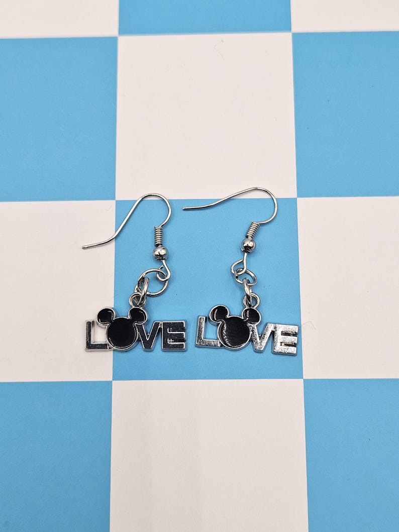 May include: Silver-toned dangle earrings. Each earring features the word "LOVE" with a black silhouette of a mouse head replacing the "O". The earrings hang from silver-colored fish hook ear wires.
