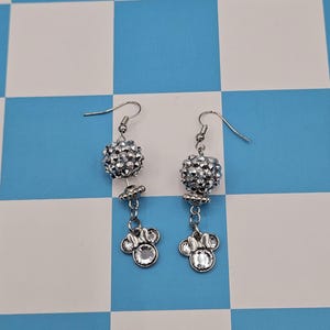 May include: A pair of silver dangle earrings. Each earring features a sparkling, round, beaded top, a small flower-shaped accent, and a charm shaped like a mouse head with a clear gem center. The earrings hang from a simple hook.