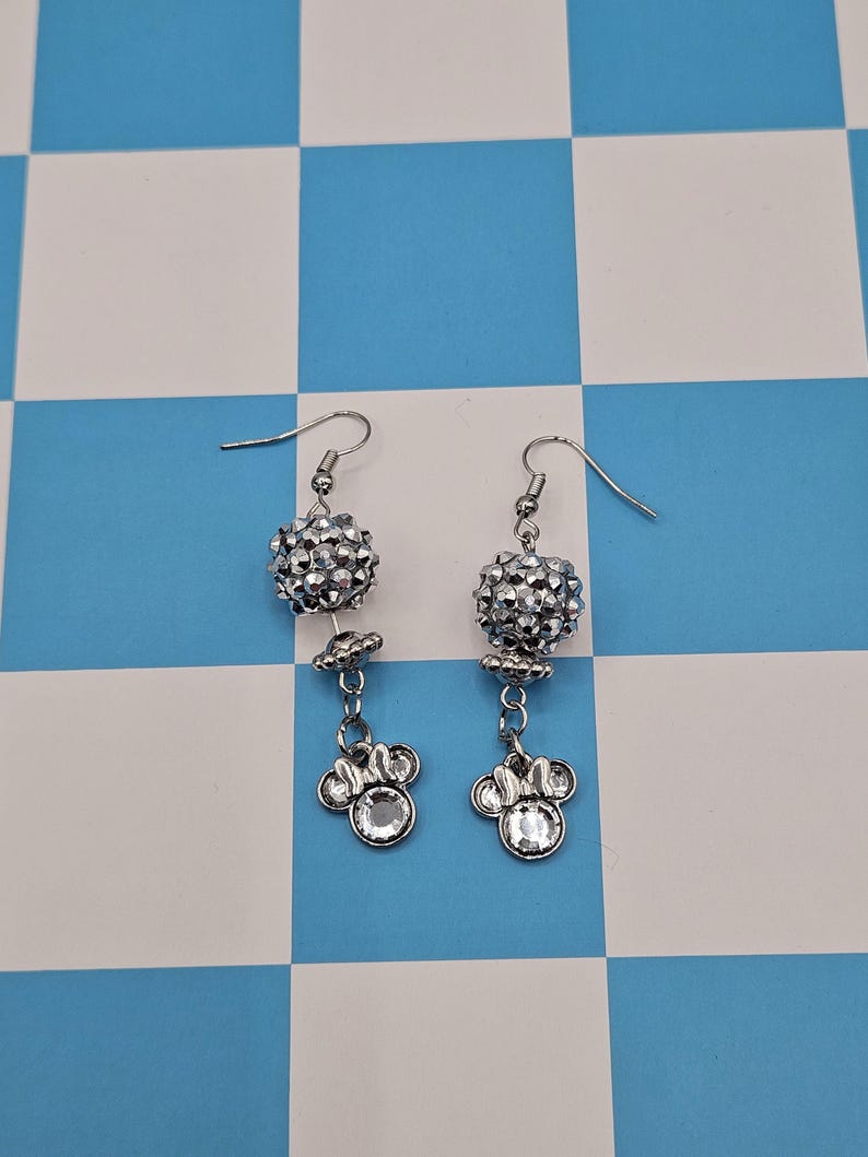 May include: A pair of silver dangle earrings. Each earring features a sparkling, round, beaded top, a small flower-shaped accent, and a charm shaped like a mouse head with a clear gem center. The earrings hang from a simple hook.