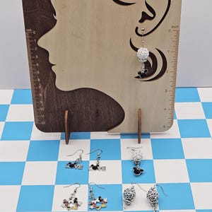 May include: Earrings displayed on a wooden stand with a profile cutout. The earrings include various designs: Mickey Mouse, the word "LOVE", and crystal-like beads. The background is a blue and white checkered pattern.