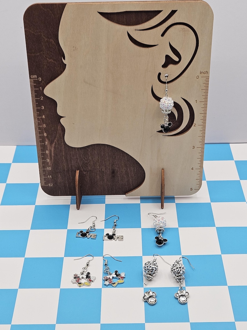 May include: Earrings displayed on a wooden stand with a profile cutout. The earrings include various designs: Mickey Mouse, the word "LOVE", and crystal-like beads. The background is a blue and white checkered pattern.