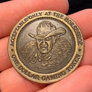 May include: A bronze one-dollar gaming token with a portrait of a man wearing a cowboy hat and fur-lined coat. The text around the edge reads "ACCEPTABLE ONLY AT THE HORSESHOE" and "ONE DOLLAR GAMING TOKEN."