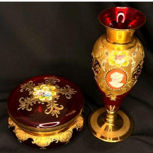 Puede incluir: Un jarrón y una caja de baratijas a juego de cristal rojo rubí. El jarrón tiene un diseño de filigrana dorada con un camafeo y detalles florales. La caja es redonda con un diseño de filigrana dorada y detalles florales. Ambas piezas tienen una base dorada.