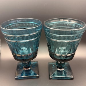 May include: Two teal-colored vintage goblets with a square base and a decorative pattern. The rims and the base of the goblets have a silver trim. The goblets are made of glass and are likely used for serving beverages.