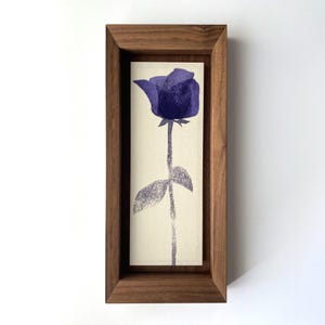 Könnte beinhalten: Ein gerahmter Druck mit einer stilisierten lila Rose. Das Kunstwerk befindet sich vor einem cremefarbenen Hintergrund und ist in einem rechteckigen, dunklen Holzrahmen eingefasst. Die Rose hat einen langen Stiel und ein einzelnes Blatt.