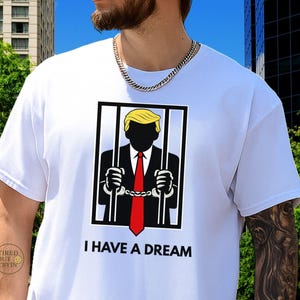 Peut inclure: T-shirt blanc avec un motif d'une personne en costume et cravate rouge derrière des barreaux, avec des cheveux jaunes. Le texte "I HAVE A DREAM" est imprimé en dessous. Le t-shirt est à manches courtes.