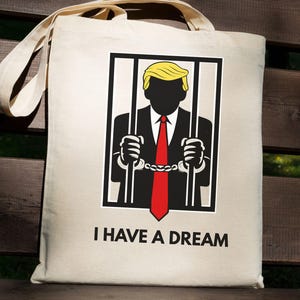May include: Beige tote bag with a graphic of a person behind bars, wearing a suit and red tie, with yellow hair. The text "I HAVE A DREAM" is printed below the graphic. The bag has long beige handles.