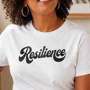 Könnte beinhalten: Ein weißes Kurzarm-T-Shirt mit dem Wort Resilience, das in einer fetten, schwarzen, kursiven Retro-Schrift auf der Brust gedruckt ist. Das Kleidungsstück wird mit einer blauen Jeans kombiniert und sorgt für einen lässigen Look mit klarem Design.
