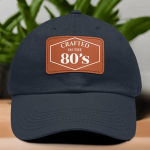 May include: Navy blue baseball cap with a brown leather patch. The patch features the text "Crafted in the 80's" in white, within a white outlined hexagon. The cap is made of a soft fabric and has a curved brim.