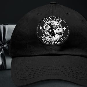 May include: Black baseball cap with a circular patch featuring a witch on a broomstick flying in front of a full moon. The patch has the text "HEX THE PATRIARCHY" in a circular design. The cap is made of a soft material.