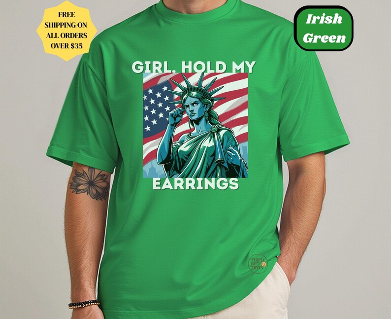 May include: Green t-shirt with a graphic of the Statue of Liberty in front of an American flag, with the text "GIRL. HOLD MY EARRINGS". The shirt also has the text "Irish Green".
