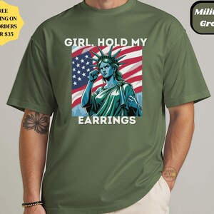 May include: Military green t-shirt featuring a graphic of the Statue of Liberty with the American flag in the background. The text reads "GIRL. HOLD MY EARRINGS". The shirt has short sleeves and a round neckline.