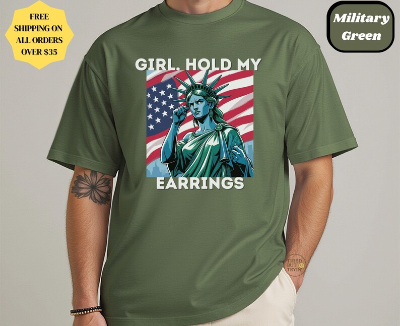 May include: Military green t-shirt featuring a graphic of the Statue of Liberty with the American flag in the background. The text reads "GIRL. HOLD MY EARRINGS". The shirt has short sleeves and a round neckline.