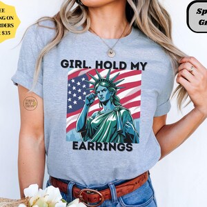 May include: Light gray t-shirt with a graphic of the Statue of Liberty in front of an American flag. The text "GIRL. HOLD MY EARRINGS" is printed on the shirt. The shirt is labeled "Sport Grey".