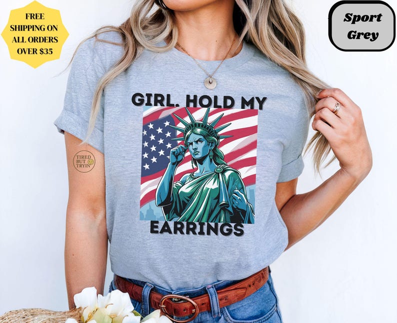 May include: Light gray t-shirt with a graphic of the Statue of Liberty in front of an American flag. The text "GIRL. HOLD MY EARRINGS" is printed on the shirt. The shirt is labeled "Sport Grey".