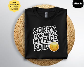 Puede incluir: Camiseta negra con el texto blanco "SORRY FOR WHAT MY FACE SAID" y un emoji amarillo con una cara triste. La camiseta se muestra sobre un salvamanteles redondo y tejido.