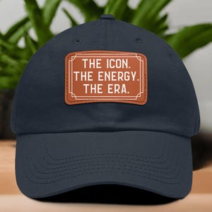 Puede incluir: Gorra de béisbol azul marino con visera curva. Un parche rectangular marrón en la parte delantera muestra el texto blanco: "THE ICON. THE ENERGY. THE ERA." La gorra está hecha de tela suave y es adecuada para el uso diario.
