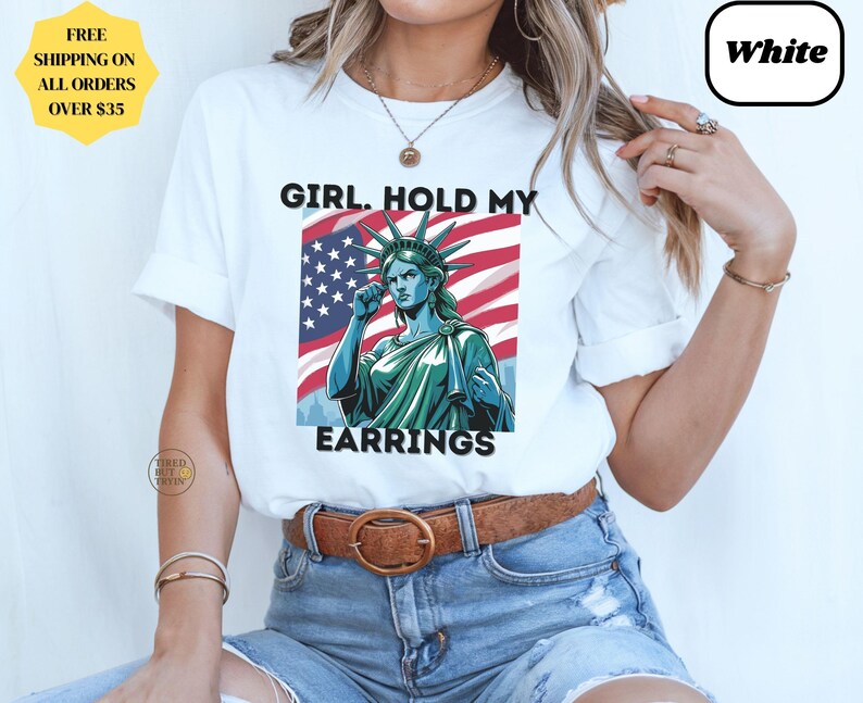 May include: White t-shirt featuring a graphic of the Statue of Liberty with the text "GIRL. HOLD MY EARRINGS" in black. The Statue of Liberty is depicted in green and blue against a backdrop of the American flag. The shirt has short sleeves.