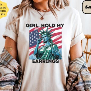 May include: A sand-colored t-shirt featuring a graphic of the Statue of Liberty with the text "GIRL. HOLD MY EARRINGS" in black. The Statue of Liberty is depicted in blue and green against a backdrop of the American flag.