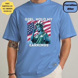 May include: Carolina blue t-shirt featuring a graphic of the Statue of Liberty with the text "GIRL. HOLD MY EARRINGS" in white. The Statue of Liberty is set against an American flag background. The shirt also has a small logo.