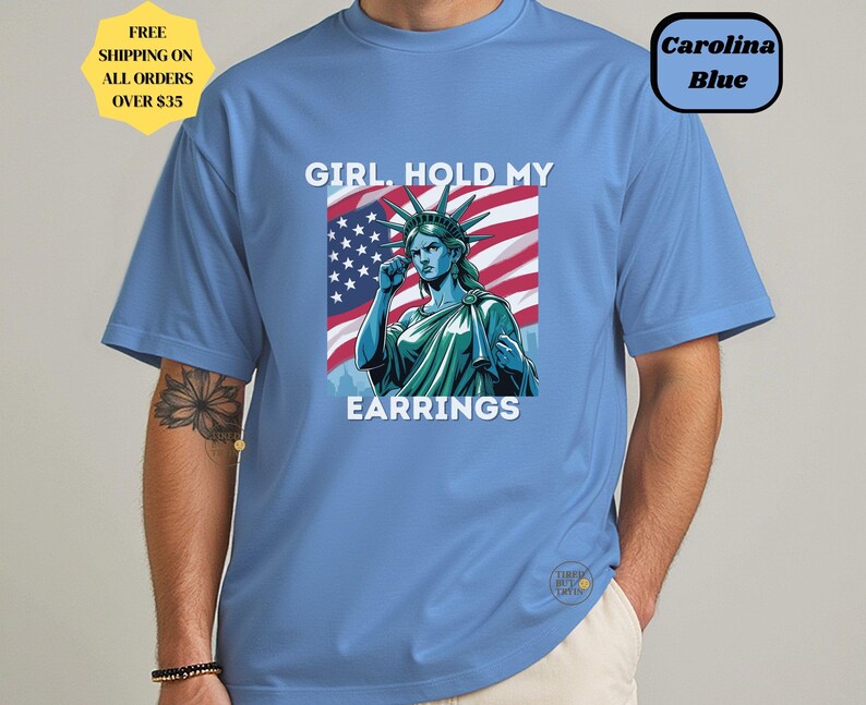 May include: Carolina blue t-shirt featuring a graphic of the Statue of Liberty with the text "GIRL. HOLD MY EARRINGS" in white. The Statue of Liberty is set against an American flag background. The shirt also has a small logo.
