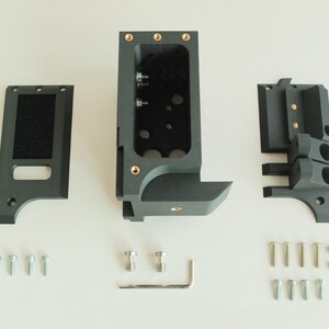 May include: An exploded view of a dark gray, 3D-printed device, likely a camera accessory or mount. The components include a main body, two side pieces, and various screws and fasteners. The parts are arranged on a white surface.
