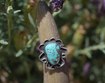May include: A silver ring with a turquoise stone. The ring features a floral design around the turquoise stone. The turquoise stone is oval-shaped with brown and blue hues. The ring is displayed on a wooden dowel against a blurred background of green and purple.
