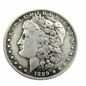 May include: A silver-toned Morgan dollar coin from 1889 featuring a profile of Lady Liberty wearing a cap inscribed with LIBERTY, surrounded by the text E PLURIBUS UNUM and thirteen stars along the circular edge of the metallic currency.