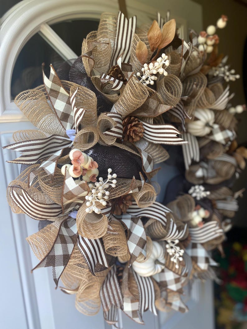 May include: A fall-themed wreath with a mix of brown, tan, and white ribbons and mesh. The wreath features a variety of textures, including burlap-like mesh, striped ribbons, and plaid ribbons. Decorative elements include small white pumpkins, pinecones, and sprigs of white berries and brown leaves.