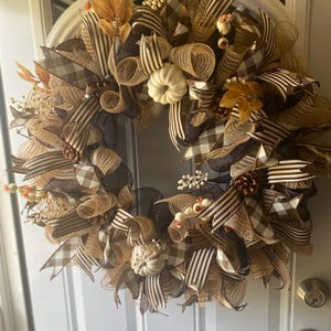 May include: A fall-themed wreath with a circular design. The wreath features a combination of brown, tan, and cream colors. It is adorned with various ribbons, including plaid and striped patterns. Decorative elements include small white pumpkins, leaves, and berries. The wreath is hanging on a white door.