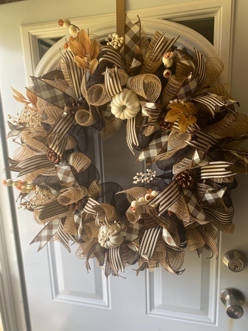 May include: A fall-themed wreath with a circular design. The wreath features a combination of brown, tan, and cream colors. It is adorned with various ribbons, including plaid and striped patterns. Decorative elements include small white pumpkins, leaves, and berries. The wreath is hanging on a white door.