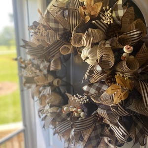 May include: A fall-themed wreath with a mix of brown, tan, and cream colors. The wreath features a variety of textures, including ribbons, mesh, and decorative elements. Accents include white pumpkins, berries, and leaves. The wreath is displayed on a white door.