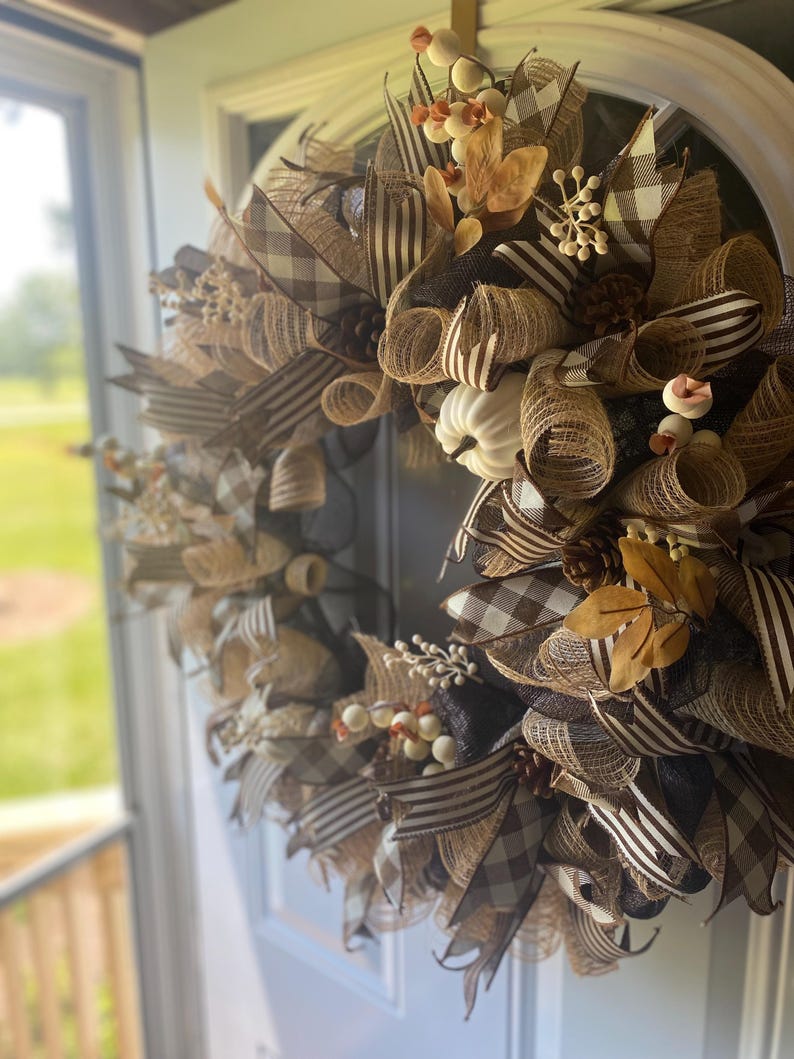 May include: A fall-themed wreath with a mix of brown, tan, and cream colors. The wreath features a variety of textures, including ribbons, mesh, and decorative elements. Accents include white pumpkins, berries, and leaves. The wreath is displayed on a white door.
