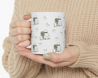 May include: White ceramic mug featuring a repeating pattern of cartoon ghosts holding flowers, bows, and hearts. The ghosts are white with black eyes, and the flowers are green and brown. The mug has a standard handle.