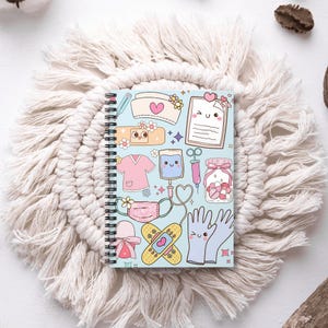 Puede incluir: Un cuaderno con espiral con una cubierta azul claro que presenta ilustraciones lindas de artículos médicos como un gorro de enfermera, vendas y una jeringa. El cuaderno tiene una encuadernación en espiral blanca.