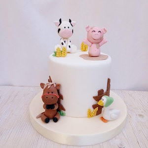 Könnte beinhalten: Eine weiße Torte, verziert mit Bauernhoftierfiguren. Die Torte zeigt eine Kuh, ein Schwein, ein Pferd und eine Ente, zusammen mit Weizenähren und einem Holzzaun. Die Torte steht auf einem weißen Tortenständer.