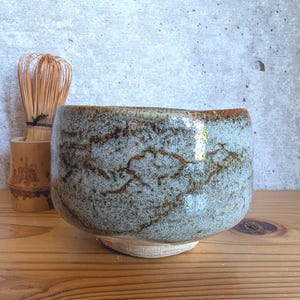 May include: A ceramic matcha bowl with a speckled light blue and grey glaze featuring dark brown crackle patterns, accompanied by a bamboo tea whisk standing in a matching bamboo holder, showcasing a textured finish and a rounded shape.
