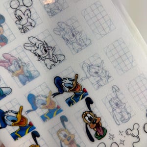 Puede incluir: Hoja de pegatinas de personajes de Disney con Mickey Mouse, Donald Duck, Pluto y otros. Algunas pegatinas son de color, mientras que otras son contornos. La hoja tiene un patrón de cuadrícula y es parcialmente transparente.