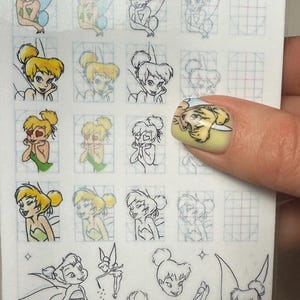 Op de afbeelding: Vel met nail art stickers met verschillende illustraties van een fee met blond haar, groene jurk en vleugels. Sommige stickers zijn gekleurd, andere zijn lijntekeningen. Een beschilderde nagel toont een bijpassend feeënontwerp.