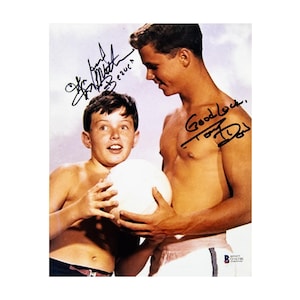 May include: A vintage color photograph featuring two shirtless individuals holding a white ball. The image includes two distinct black ink signatures and the handwritten text Good luck. The individuals are positioned against a light sky background.