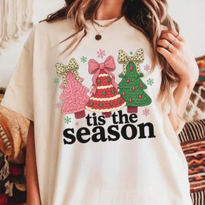 Puede incluir: Camiseta color crema con tres árboles de Navidad en rosa, rojo y verde, cada uno adornado con un lazo. El texto "tis the season" está impreso en negro debajo, con copos de nieve alrededor del diseño.