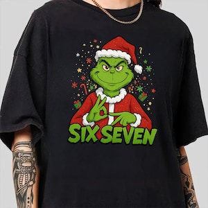 Puede incluir: Camiseta negra con un personaje de dibujos animados verde con gorro de Papá Noel y traje rojo, haciendo un gesto con la mano. El texto "SIX SEVEN" está impreso en verde debajo. Estrellas y copos de nieve festivos rodean al personaje.