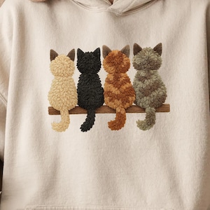 Könnte beinhalten: Beige Kapuzenpullover mit einem gestickten Design von vier Katzen, die auf einem Holzast sitzen. Die Katzen sind in den Farben Beige, Schwarz, Orange und Grün dargestellt. Der Kapuzenpullover hat eine neutrale Farbe.