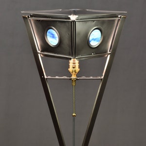 May include: A unique, geometric table lamp with a dark metal frame and a square base. The lamp features a triangular top with two circular windows displaying a blue sky design. The lamp has a brass-colored bulb socket and a pull chain.