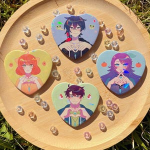 May include: Four heart-shaped anime character pins on a wooden tray, each featuring a different character design. The pins are surrounded by small, clear, decorative crystals. The website address hynorin.etsy.com is visible at the bottom.