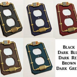 May include: Six different colored outlet covers with a steampunk design. The covers are black, dark blue, dark red, brown, and dark green. The covers have a gold trim and a gold coil on the bottom. The covers are all in a rectangular shape.