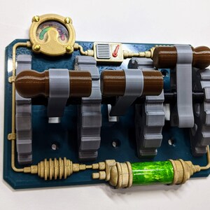 May include: A steampunk-style light switch plate with three toggle switches, each with a brown knob and a gray gear. The plate is painted a dark teal blue and features gold accents, including a gauge with a rainbow needle and a green glass tube.